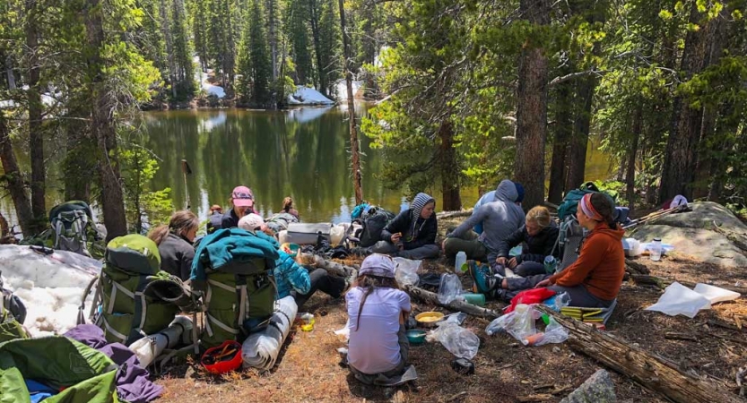 A group of people rest near a body of water in a wooded area. 